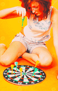 Woman listlessly dropping darts onto a dartboard lying on the ground.